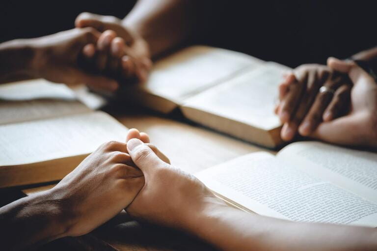 Christian group of people holding hands praying worship to believe and Bible on a wooden table for devotional or prayer meeting concept.